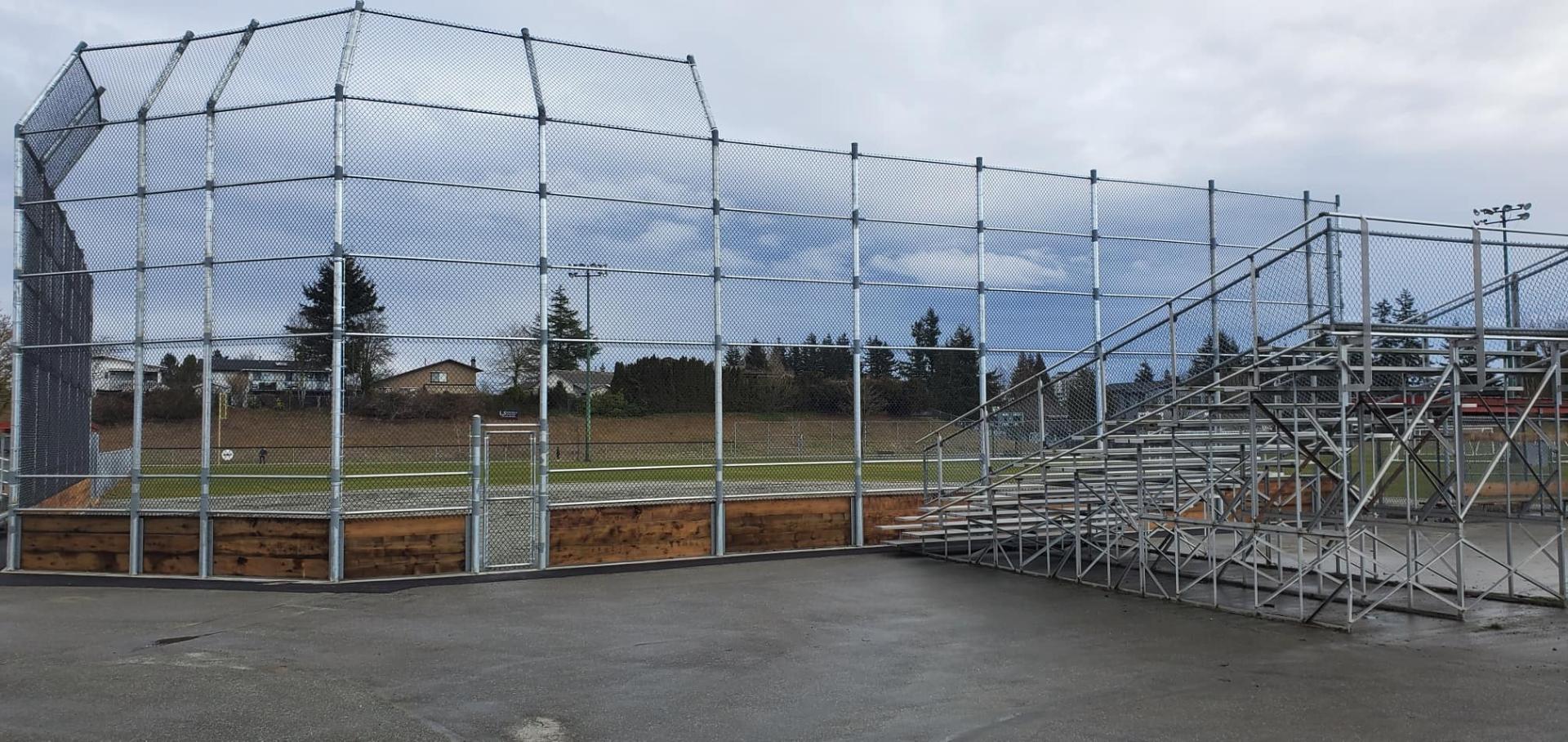 Tall galvanized chain link baseball backstop with overhang and cedar kickboard next to bleachers — Richmond, BC commercial project.