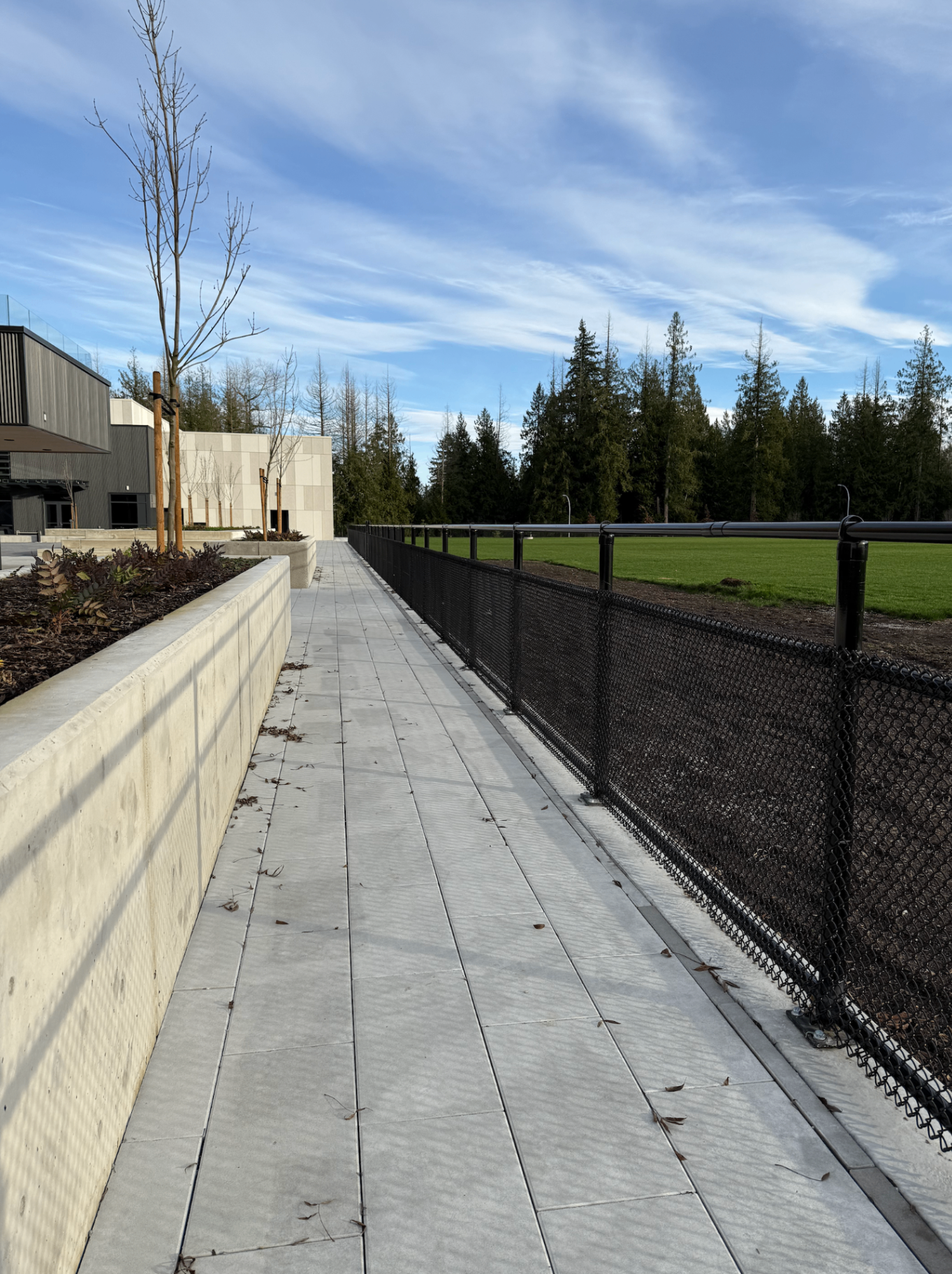 Black chain link fence with galvanized pipe top railing lining a concrete walkway at a government sports facility in Langley, BC.
